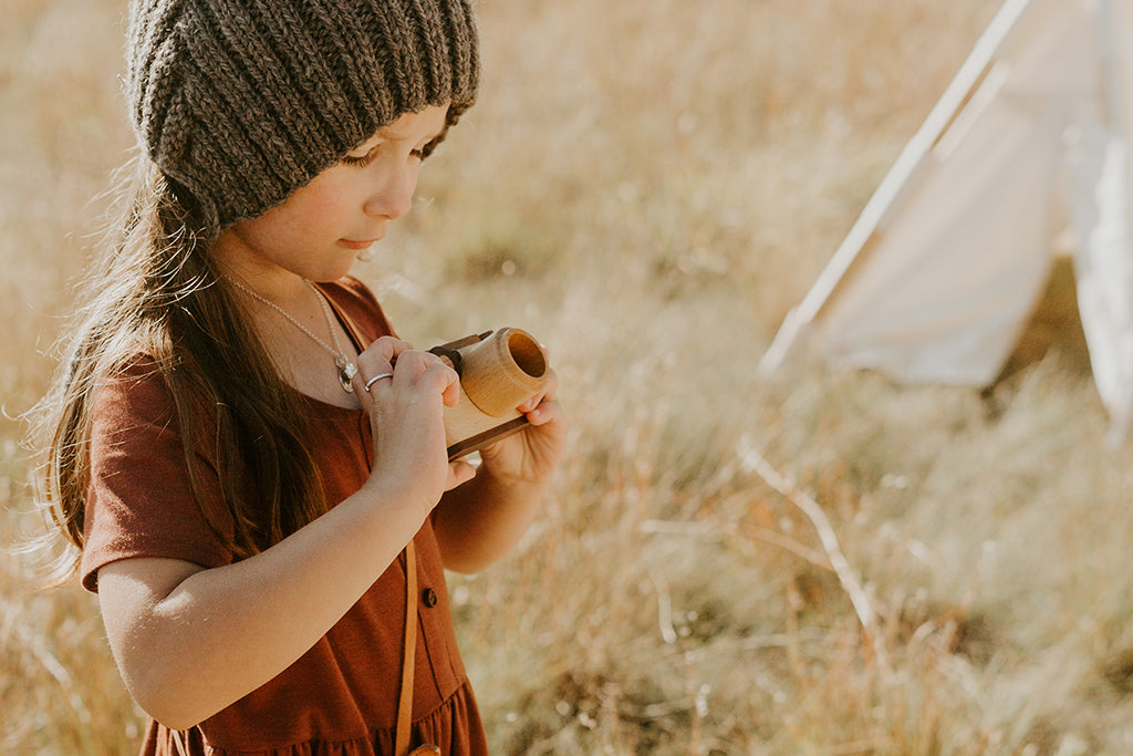 A young girl wearing a knit hat and fall colored dress holds a wooden toy camera outdoors, pretend-taking photos in a sunny field.