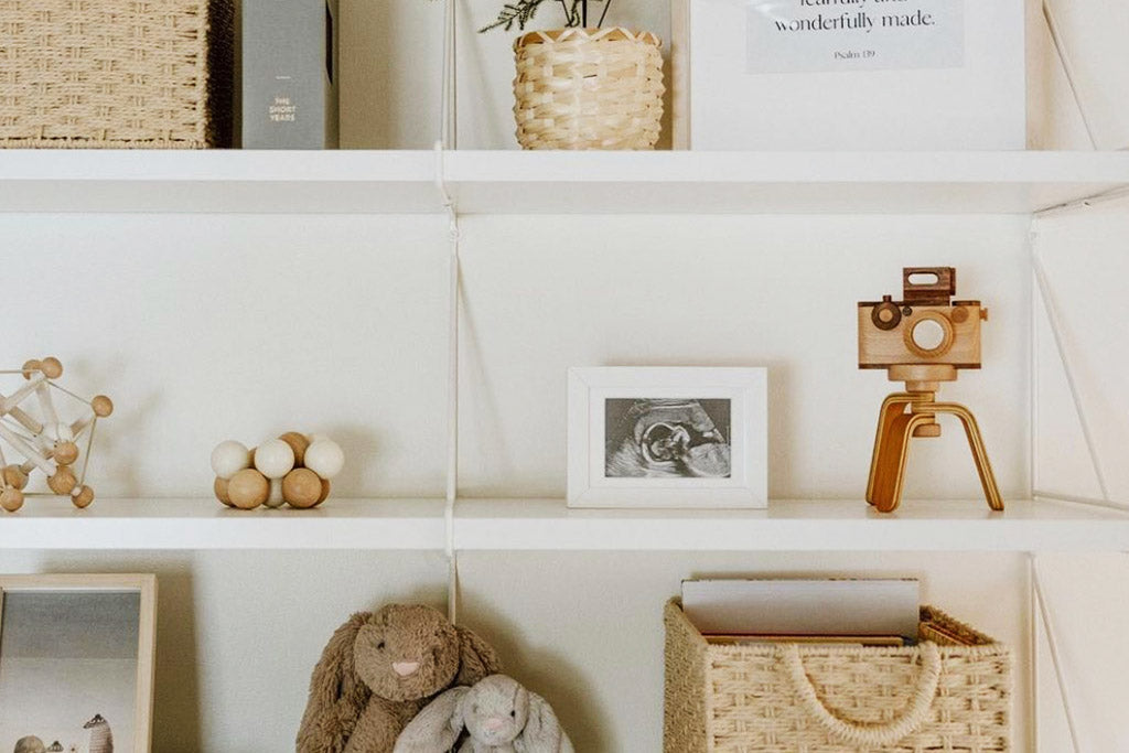 A wooden toy camera with a kaleidoscopic lens sits on a minimalist white shelf, styled as decor alongside woven baskets, soft toys, and a framed ultrasound photo.