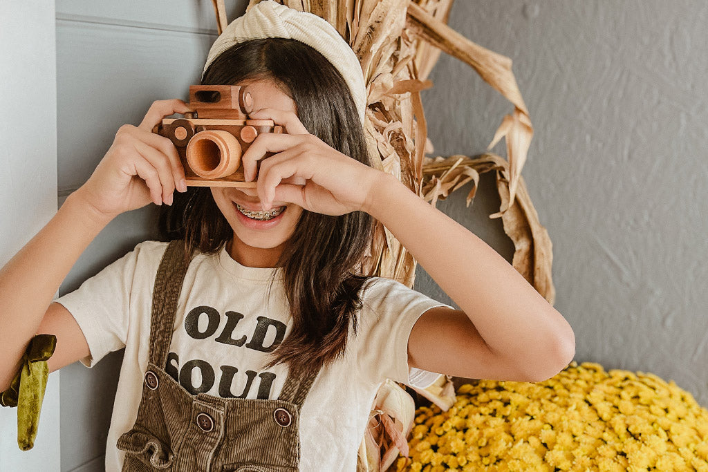 Child holding a wooden toy camera with a big smile, pretending to be a photographer; vintage-inspired 35MM camera design with a flash, made from non-toxic and biodegradable materials, encouraging creativity and open-ended play.
