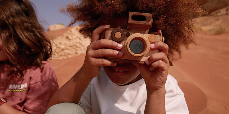 The photo shows a boy holding a wooden toy camera pretend to play photographer. He is playing his toy camera outdoor taking photo of his model. 