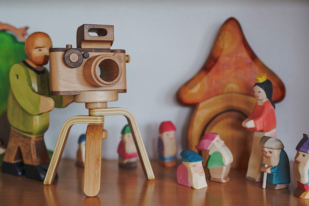 A wooden doll pretends to take a photo with a wooden toy camera on a tripod, featuring a vintage-inspired 35MM design and flash, made from non-toxic, biodegradable materials, perfect for open-ended play and fostering creativity.