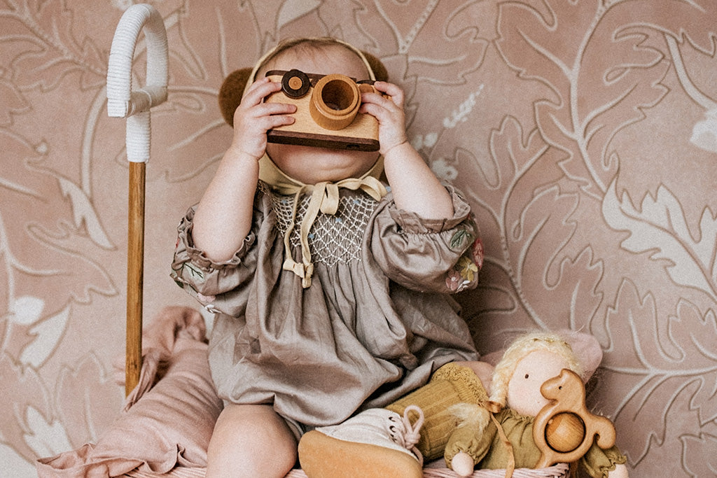 Toddler in a vintage-style dress looking through the kaleidoscope lens, pretending to take a photo with a wooden toy camera.