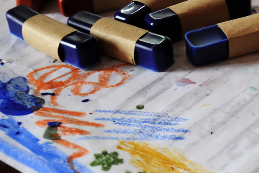 Dark blue non-toxic beeswax oil pastels with kraft paper labels beside a colorful eco-friendly drawing on white paper