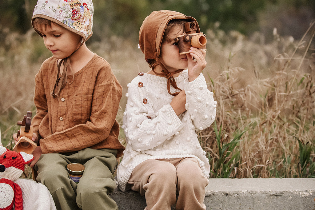 Two children dressed in cozy clothing, sitting outdoors, one pretend-taking photos with a wooden toy camera that has a vintage-inspired design, featuring a kaleidoscope lens and clickable buttons.