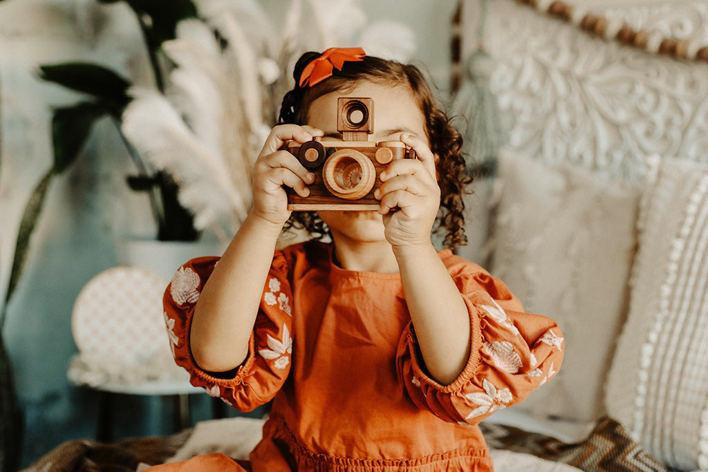A toddler girl playing a wooden toy camera with a vintage-inspired design, featuring a kaleidoscope lens and detachable flash, perfect for children's pretend play.