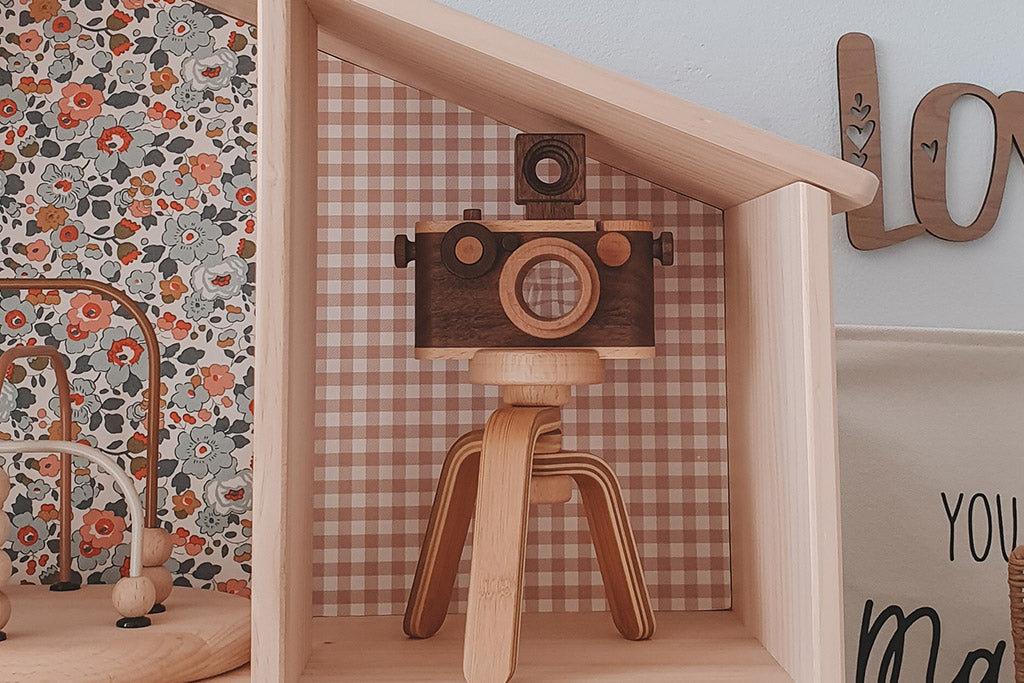 A vintage-inspired wooden toy camera with a kaleidoscope lens, placed on a wooden tripod, displayed on a gingham-backed shelf surrounded by playful decor.
