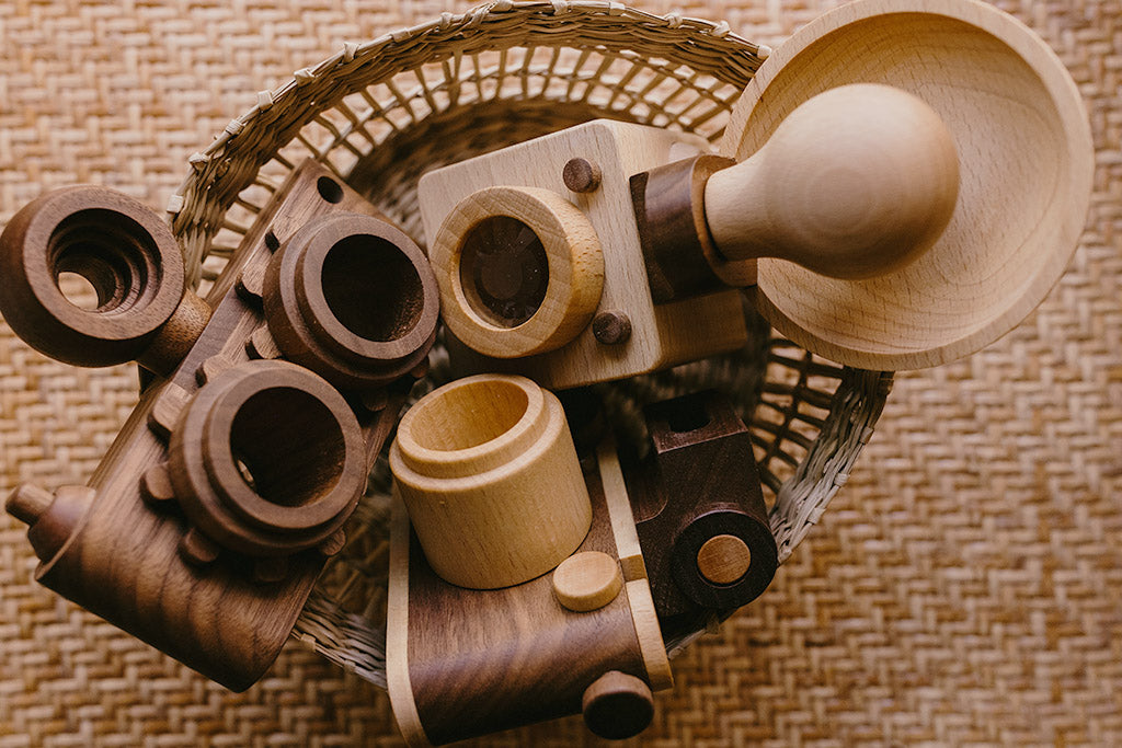 A basket filled with handcrafted wooden toy cameras, including a Brownie-inspired camera with a bulb-style flash, made from high-quality beechwood and walnut.