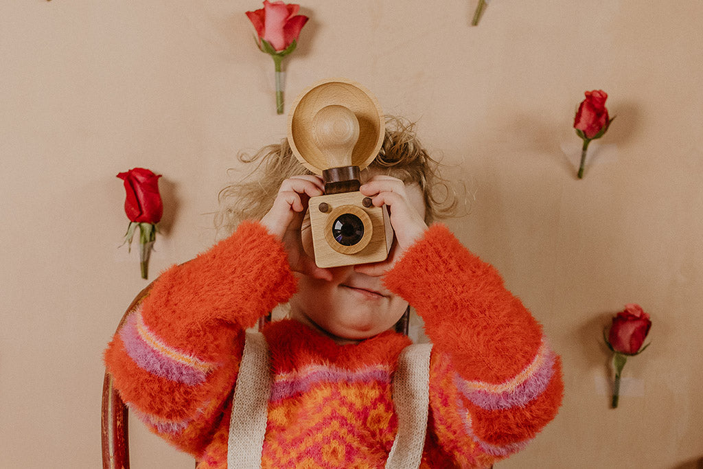 A toddler girl in a red sweater looks through a high-quality beechwood Brownie-inspired wooden toy camera. This modern take on a classic design is non-toxic, biodegradable, and ideal for children’s open-ended playtime.