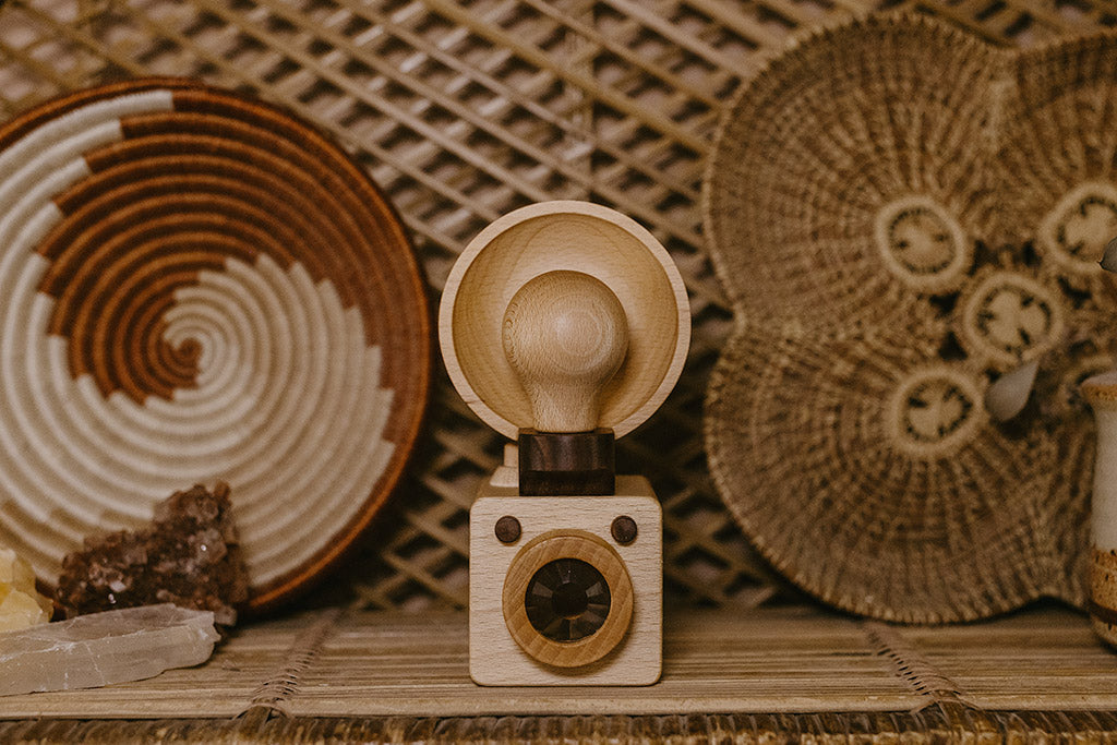 The Brownie wooden toy camera is inspired by the vintage Kodak Brownie camera featuring a detachable bulb-style flash and kaleidoscope lens, displayed against a backdrop of woven and earthy decor elements.