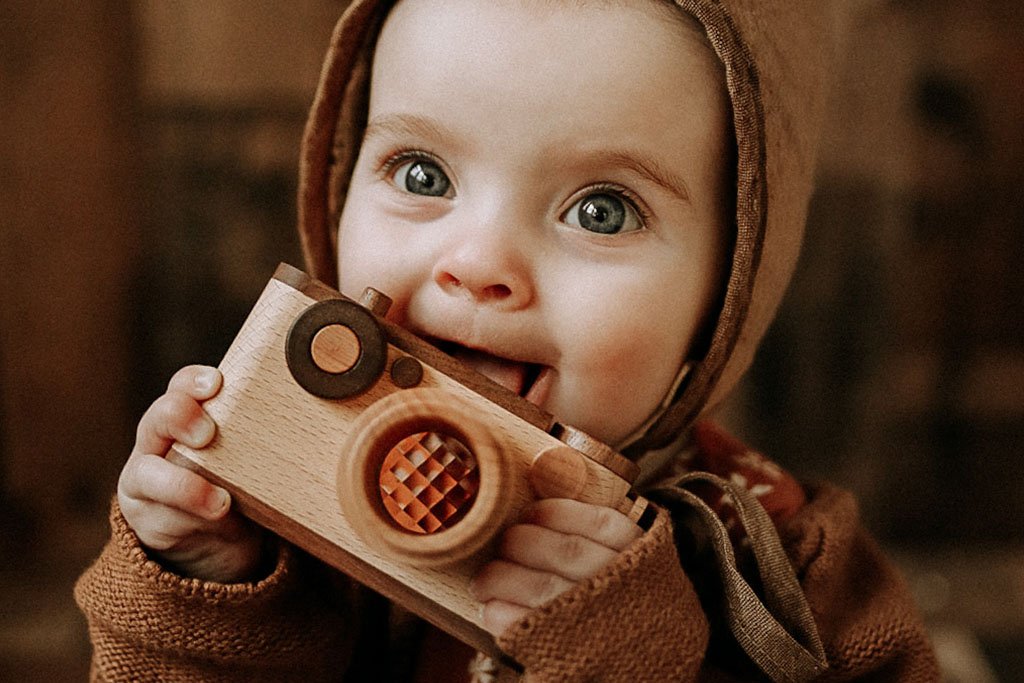 A baby with bright eyes holds a wooden toy camera with a kaleidoscopic lens, exploring it with curiosity, showcasing its safe, child-friendly design and sustainable materials.