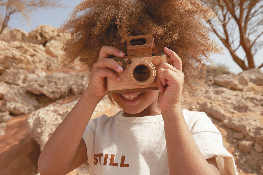 A wooden toy camera with a kaleidoscopic lens and vintage-inspired design is held by a smiling child outdoors, pretending to take photos, showcasing its playful and creative features of the camera.