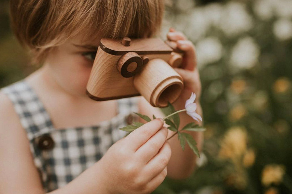 A wooden toy camera with a vintage-inspired design is held by a child outdoors, zooming in on a wild flower, showcasing its playful and creative potential for pretend play.