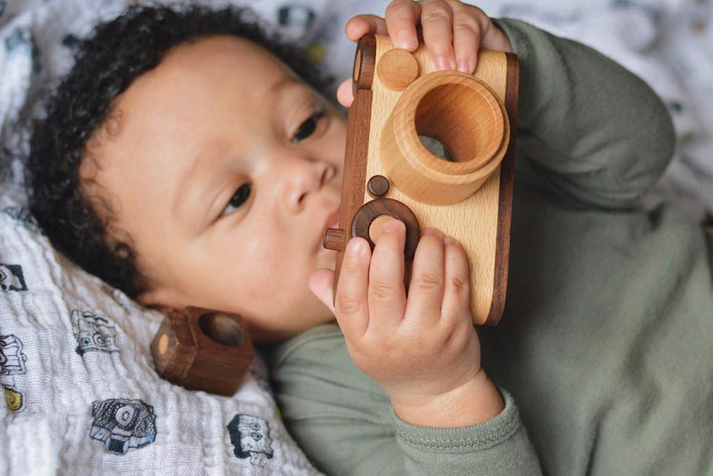 A baby boy holding a wooden toy camera with a kaleidoscope lens and vintage-style flash, pretend-playing as a photographer. Crafted from non-toxic, biodegradable materials, it encourages creativity and open-ended playtime.