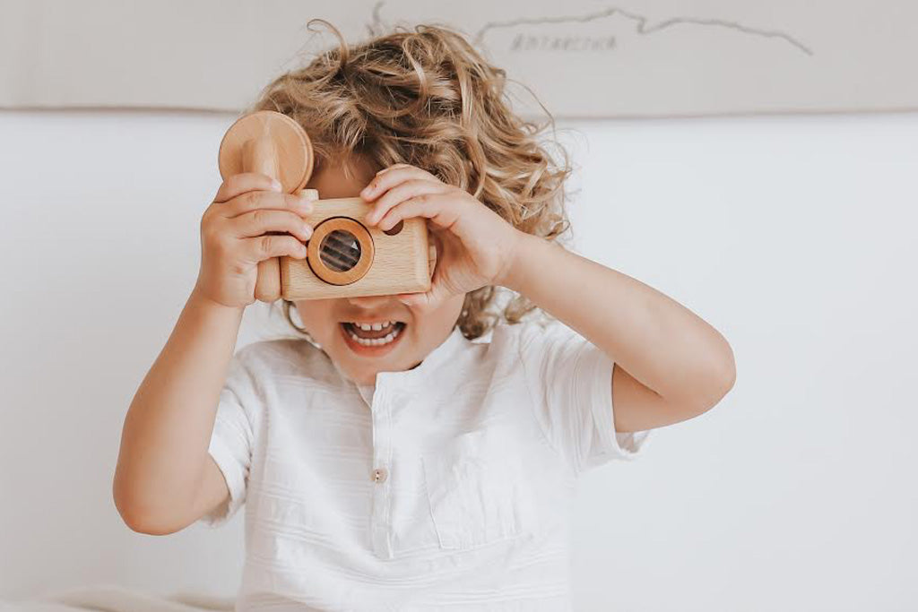 Photo shows a child holding wooden toy camera with a big smile behind the camera, he is pretend-playing photographer. Inspired by the vintage 35MM camera, the toy camera comes with a vintage flash, and it’s the best first camera for encouraging children’s creativity. Wooden toys are safer than plastic toys because they are made of non-toxic and biodegradable materials. When choosing wooden toys, wooden toy camera is the essential type of toy for your child’s open-ended playtime.