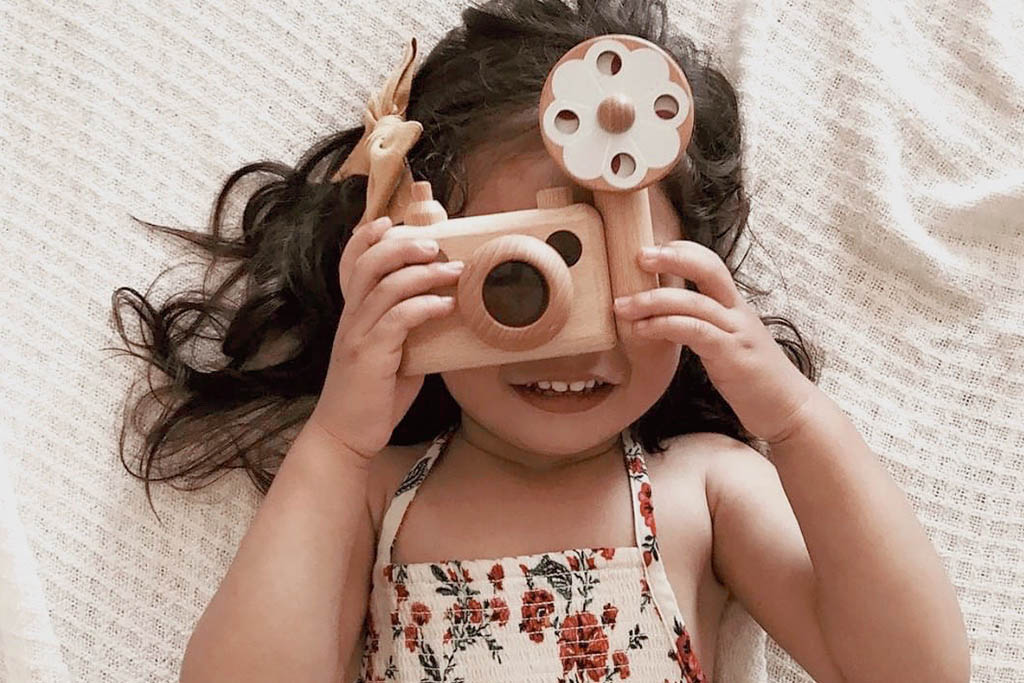 Photo shows a child holding wooden toy camera with a big smile behind the camera, she is pretend-playing photographer. Inspired by the vintage 35MM camera, the toy camera comes with a vintage flash, and it’s the best first camera for encouraging children’s creativity. Wooden toys are safer than plastic toys because they are made of non-toxic and biodegradable materials. When choosing wooden toys, wooden toy camera is the essential type of toy for your child’s open-ended playtime.