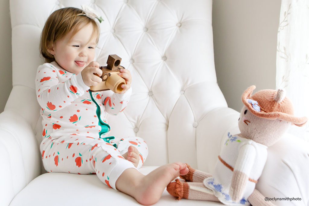 Toddler in floral pajamas pretend play as a photographer with a wooden toy camera, taking photos of a knitted doll on a white chair.