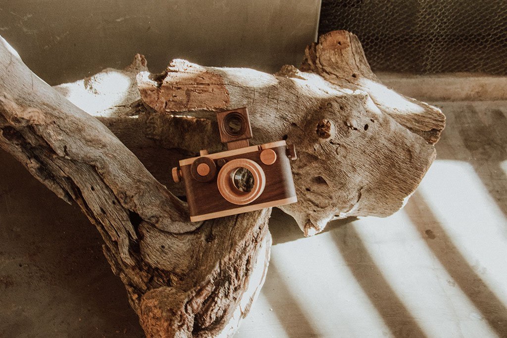 A vintage-inspired wooden toy camera with a kaleidoscope lens and detachable flash, placed on rustic driftwood in warm, natural lighting.