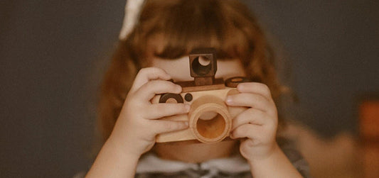 photo shows a girl holding a wooden toy camera pretend play as a photographer taking photo of her mama