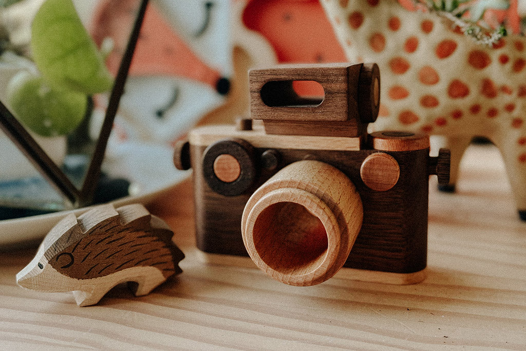 A vintage-inspired wooden toy camera with a detachable flash and clickable button, displayed on a wooden surface alongside a small wooden hedgehog figurine, surrounded by playful and natural decor elements.