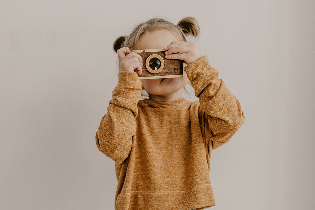 Child holding a 35MM-style wooden toy camera with kaleidoscopic lens, flash, and button; eco-friendly and heirloom quality, ideal for pretend play and sensory exploration.