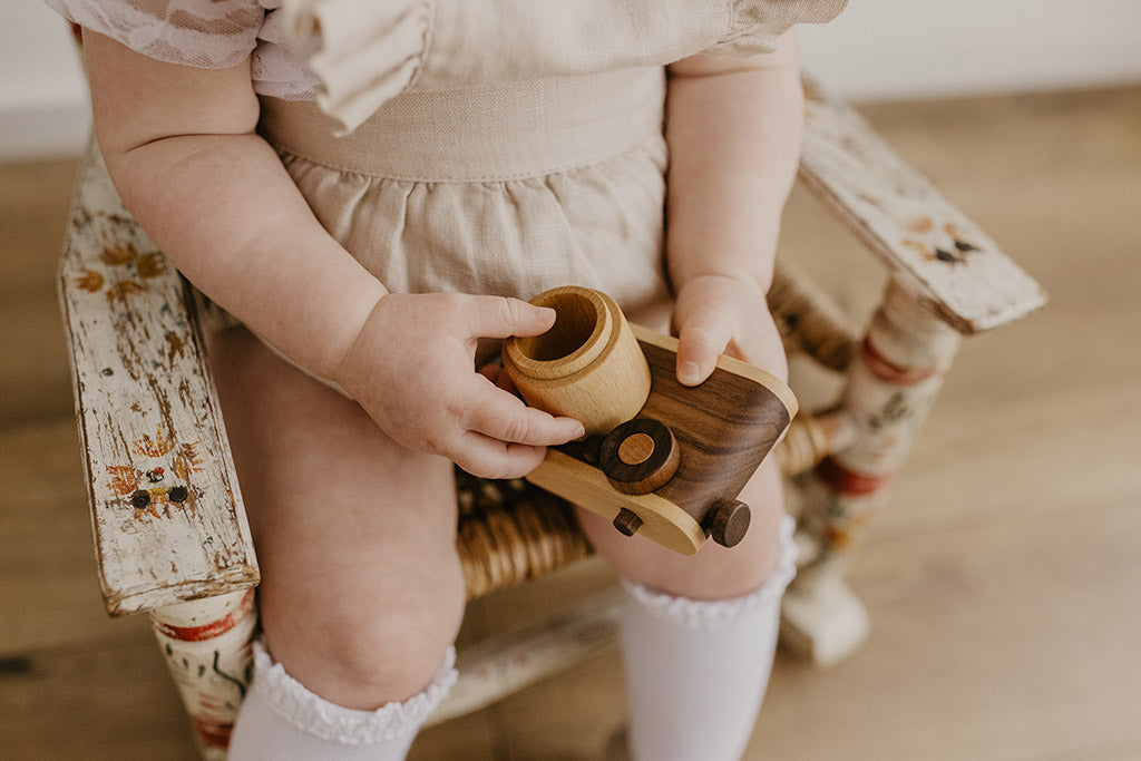 A toddler seated on a vintage wooden chair holds a wooden toy camera with a kaleidoscopic lens, designed for open-ended play.