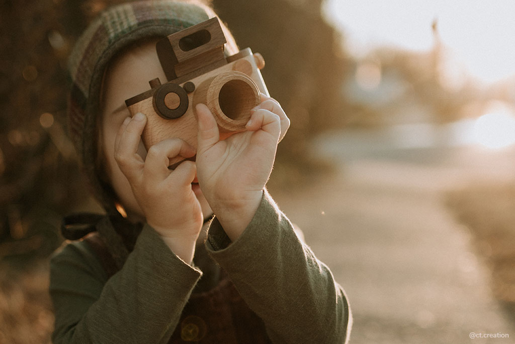 Father's Factory wooden toy camera, 35MM Original wooden toy camera with detachable magnetic flash, clickable button, and kaleidoscopic lens. It’s the perfect toy camera for pretend play, sensory play, and homeschooling. This camera for kids is made of walnut and beechwood with heirloom quality. Photo shows a toddler holding a wooden toy camera, and pretend to take photos. 